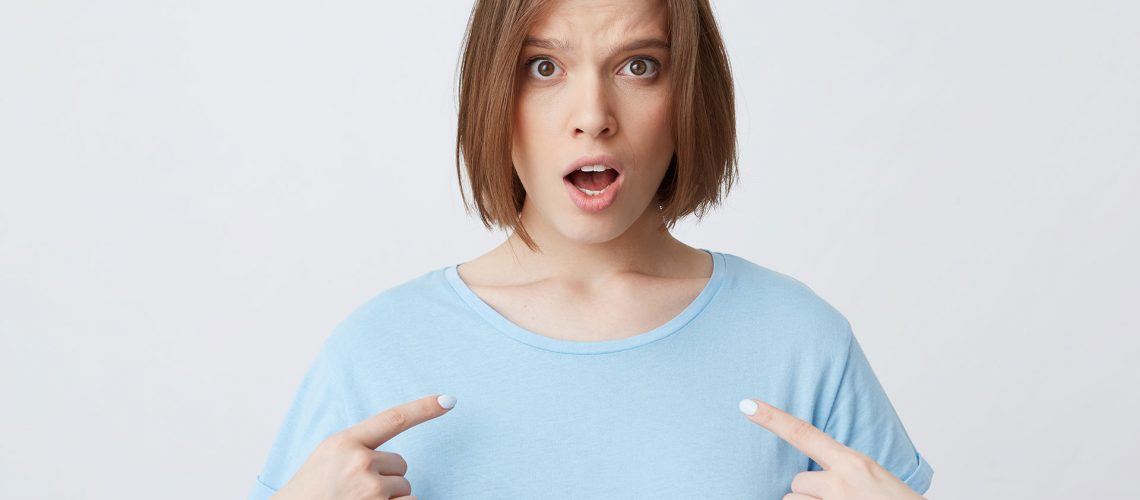 Amazed shocked young woman in blue t shirt with opened mouth looks stunned and points at herself with fingers on both hands isolated over white background